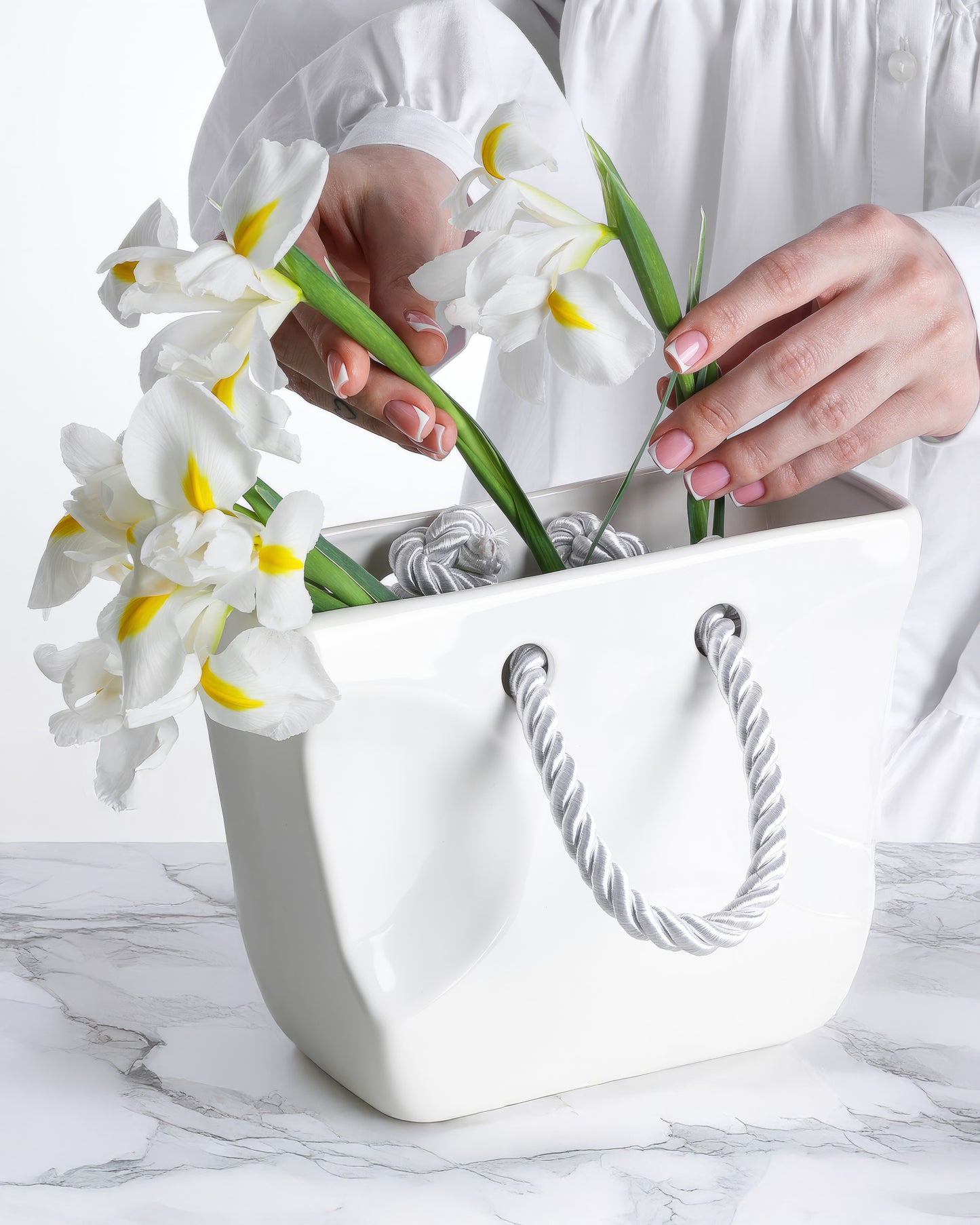Hands arranging white irises in a white ceramic purse vase with rope handles, gift for her home decor